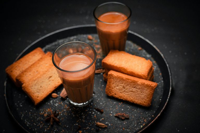 Two glasses of steaming masala chai with whole spices and ginger on a wooden tray in an Indian kitchen
