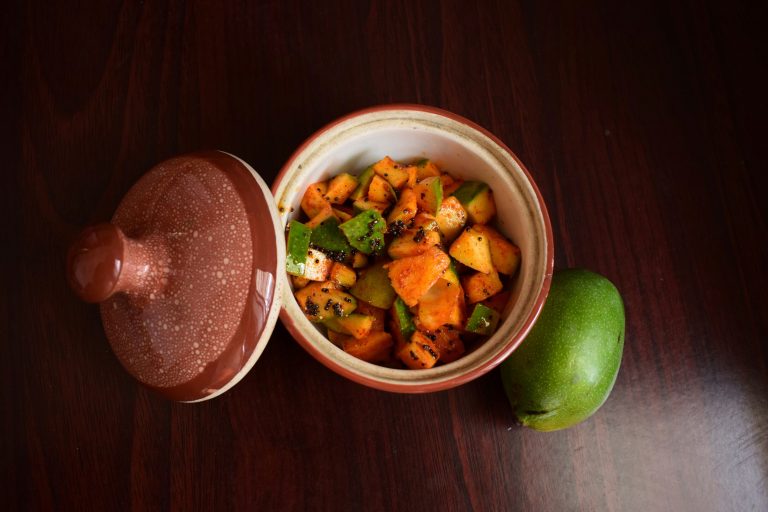 Glass jar of homemade raw mango pickle with whole spices and mustard oil on a rustic wooden surface