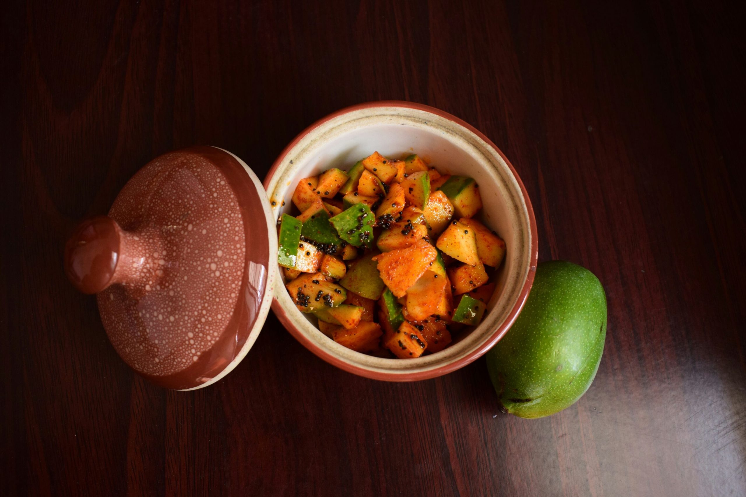 Glass jar of homemade raw mango pickle with whole spices and mustard oil on a rustic wooden surface