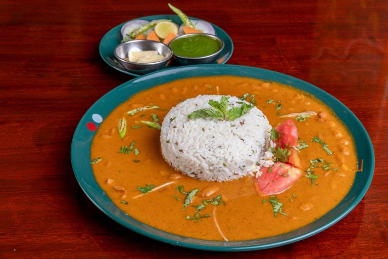 A bowl of deep red rajma curry with steamed white rice, sliced onions and a wedge of lime on a steel thali